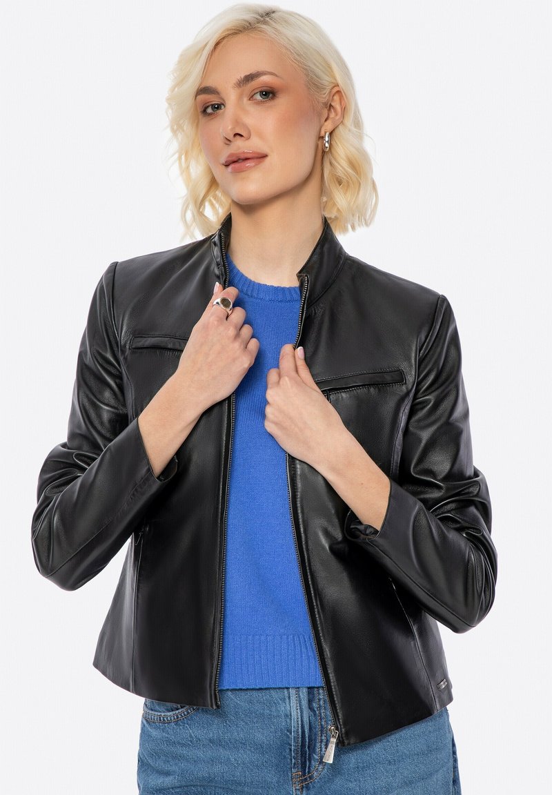 Schwarze Lederjacke mit Stehkragen, Reißverschluss vorne und flachen Brusttaschen. Trägt sie über einem blauen Pullover, kombiniert mit Jeans. Glatte Textur.