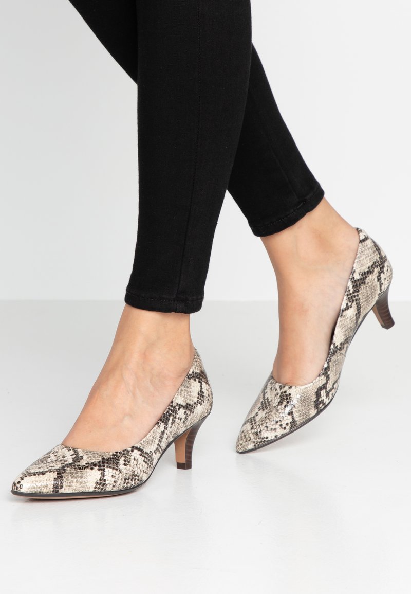 Pieds portant des chaussures à bout pointu et talons moyens avec un motif beige et marron de peau de serpent, associés à un pantalon noir ajusté.