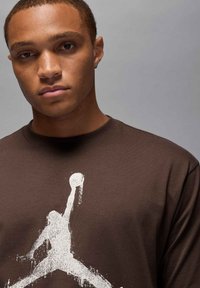 Camiseta de algodón marrón con un gráfico blanco de un atleta saltando sosteniendo un baloncesto. Diseño de cuello redondo, textura suave, ajuste casual.