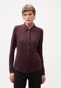 Chemise bordeaux à manches longues avec un col classique, fermeture par boutons sur le devant et un léger éclat. Présente une coupe ajustée et une texture lisse.