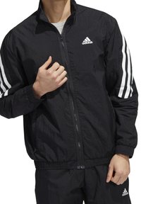 Svart vindjacka med dragkedja framtill, hög krage och elastiska manschetter. Tre vita ränder på ärmarna och en Adidas-logotyp på bröstet.