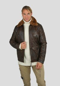 Veste en cuir marron avec un col en fausse fourrure, dotée d'une fermeture éclair et de deux poches avant. Le matériau a une finition texturée. Un pull en maille léger est porté en dessous.