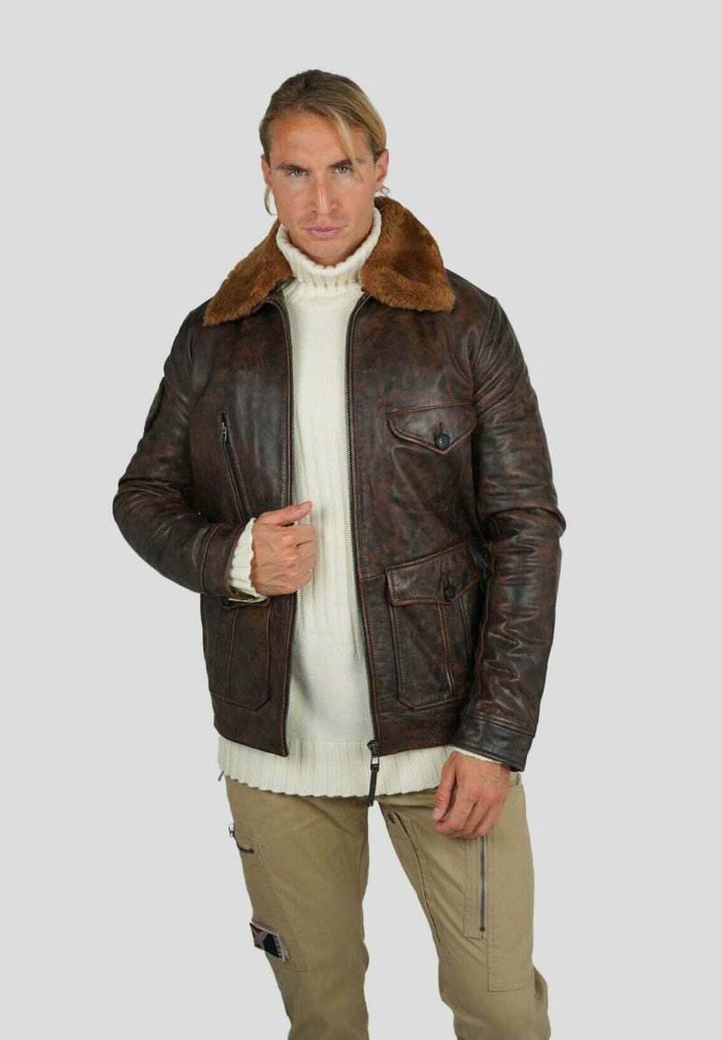 Veste en cuir marron avec un col en fausse fourrure, dotée d'une fermeture éclair et de deux poches avant. Le matériau a une finition texturée. Un pull en maille léger est porté en dessous.