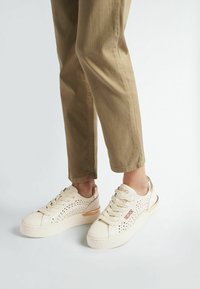 Sneakers bianche con design perforato, dettagli in marrone e suole spesse. Presentano lacci piatti e un logo sul lato e sul tallone.