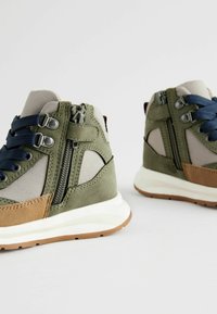 Baskets montantes vertes et brunes avec des panneaux en mesh, des lacets navy, des fermetures éclair sur le côté et des semelles en caoutchouc. Présente un design texturé et des accents contrastés.