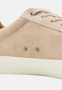 Beige velúrbőr sneaker, fehér gumitalppal, finom perforációkkal az oldalán és b�ézs fűzőkkel. Sima textúra és egyszerű dizájn.