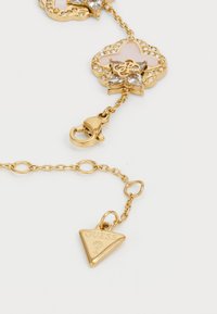 Gold-colored chain bracelet featuring a floral pendant adorned with clear rhinestones and a triangle charm engraved with "GUESS."