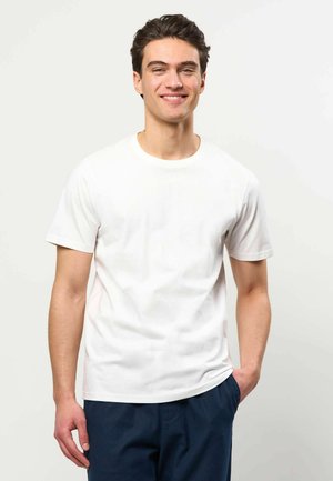 Unge mand med mørkt hår smiler, iført en ensfarvet hvid kortærmet t-shirt og marineblå bukser, stående mod en lys baggrund.