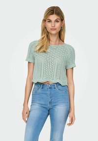 Lys grøn, strikket crop top med korte ærmer, der har et indviklet blondermønster og bølget kant, parret med blå højtaljede jeans.