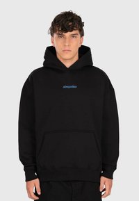 Young man with curly hair wearing a black hoodie with a front pocket and small blue "dropsize" logo on the chest, standing neutral.