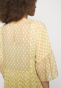 Żółta sukienka w wzory z geometrycznym motywem, krótkimi bufiastymi rękawami i dekoltem w kształcie łezki z zapięciem na guzik. Gładki, lekki materiał.