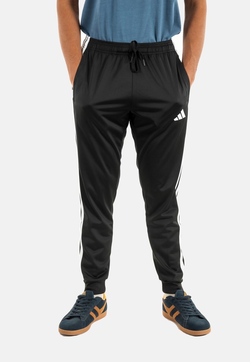 Pantalons de sport noirs avec des bandes blanches sur les côtés, taille élastique et cordon de serrage. Portés avec des chaussures navy avec des accents beige.