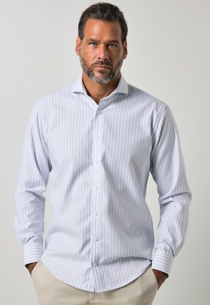 Man met baard die een lichtblauw-wit gestreept overhemd met lange mouwen en beige broek draagt, staat met handen in de zakken tegen een effen achtergrond.