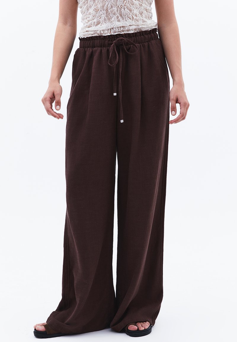 Široké hnědé kalhoty vyrobené z lehké tkaniny, s elastickým pasem na šňůrku a kontrastními bílými akcenty.