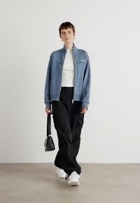 Light blue zip-up jacket, white top, black cargo pants, white sneakers, holding a small black bag. Smooth fabric, relaxed fit, minimal design.