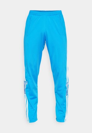 Jasnoniebieskie spodnie sportowe z białymi paskami po bokach i kieszeniami na suwak, z naszywkami z logo Adidas na każdej nogawce.