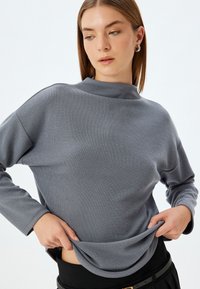Maglia grigia a maniche lunghe con collo alto e vestibilità rilassata. Tessuto morbido, materiale leggero e motivo discreto. Vita nera visibile nella parte inferiore.
