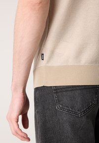 Beige kötött pulóver dombornyomott mintával, bordás aljjal és kis fekete logóval; sötét farmerrel párosítva, hátizsebének varrásával.