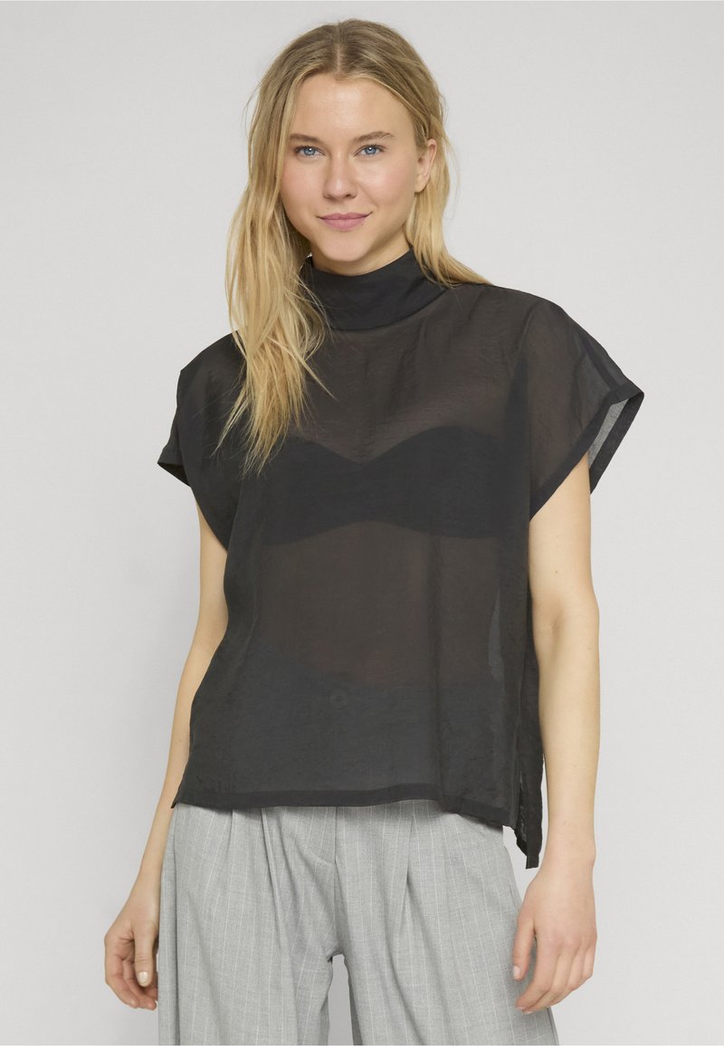 Femme blonde portant un haut noir transparent à manches courtes par-dessus un soutien-gorge noir et un pantalon large gris à fines rayures, debout devant un fond uni.