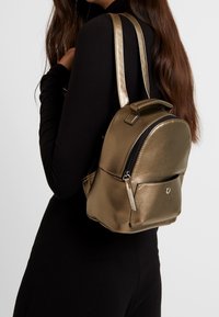 Metallisch goldener Rucksack mit runder Form und glatter Textur. Verfügt über einen oberen Griff und eine Vordertasche mit Reißverschluss sowie silbernen Hardware-Akzenten.