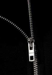 Primo piano di un indumento nero con cerniera metallica argentata e cursore marchiato con la scritta "WEEKDAY". La superficie è liscia.