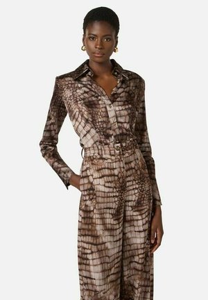 Donna che indossa una camicia a maniche lunghe marrone e beige con motivo a coccodrillo e pantaloni a vita alta abbinati, posando con una mano in tasca.
