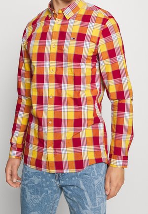 Hombre con camisa de manga larga abotonada con patrón de cuadros en rojo, naranja y amarillo y vaqueros azul claro con estampado abstracto.