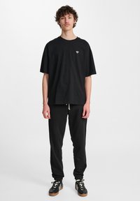 Camiseta de algodón negra con cuello redondo, corte holgado y pequeño gráfico en el pecho, combinada con pantalones de chándal negros y zapatillas negras.