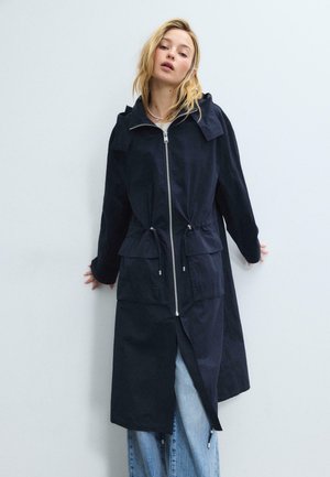 Kvinde iført en lang sort frakke med lynlås, forlommer og snore, over lyseblå jeans, stående mod en ensfarvet hvid væg.