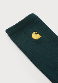 Mørk teal strikket sokk med ribbet tekstur, og med en fremtredende gul brodert logo nær toppen.
