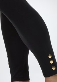 Detaljni prikaz crnih tajica s tri zlatna gumba blizu gležnja na bijeloj pozadini.