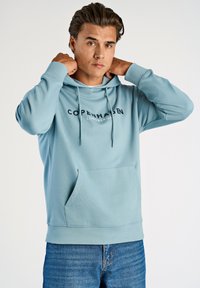Sudadera azul claro con cordón ajustable, bolsillo frontal y logo de texto "Copenhague". Tejido suave con puños y dobladillo acanalados.