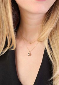 Gouden ketting met een gelaagde schakelontwerp; bevat een ronde hanger met een parel, gedragen op een blootgestelde nek. Donkere kleding contrasteert met het goud.
