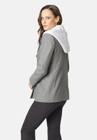 Mujer con cabello castaño largo que lleva un blazer gris de rayas con una capucha gris claro y pantalones negros, mirando por encima del hombro.