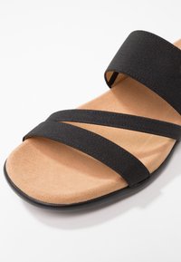 Sandales en tissu noir avec deux lanières croisées, une semelle intérieure rembourrée couleur tan et une semelle en caoutchouc noir. La texture de la surface est lisse.