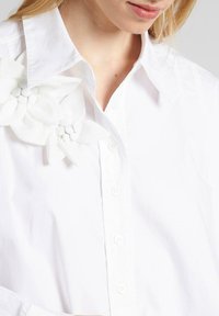 Camisa blanca de botones con un gran adorno floral de tela texturizada en el collar. Material de algodón suave, diseño clásico.