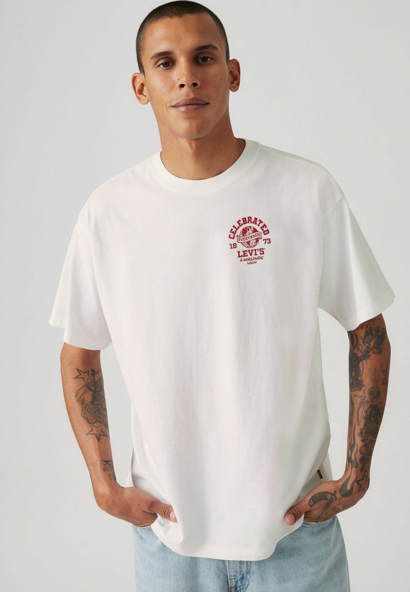 Mand med tatoveringer iført en hvid Levi's t-shirt med rødt logotekst på brystet, står med hænderne i lommerne på lyseblå jeans mod en ensfarvet baggrund.