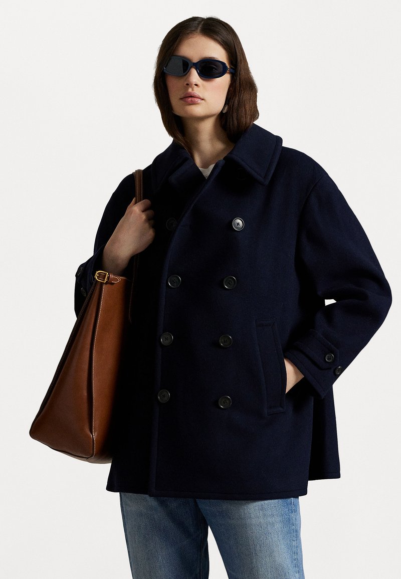 Marineblauwe wollen peacoat met dubbelknoopsluiting, grote kraag, zijzakken, gecombineerd met een bruin leren tas en donkergekleurde zonnebril.