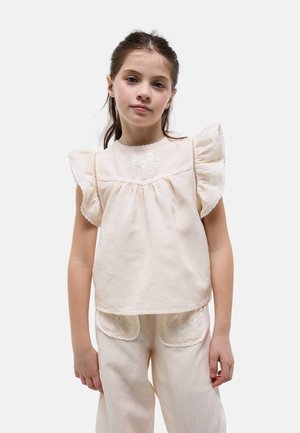 Jeune fille aux cheveux bruns portant un chemisier crème à manches à volants et un pantalon assorti avec des poches bordées de dentelle, debout devant un fond uni.