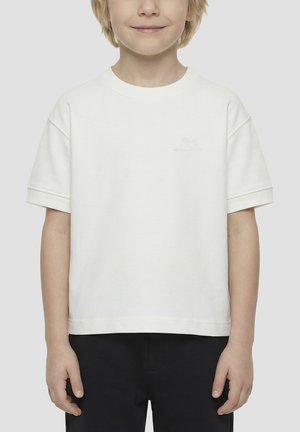 Enfant portant un t-shirt blanc uni à manches courtes et un pantalon noir, debout devant un fond neutre, souriant légèrement.