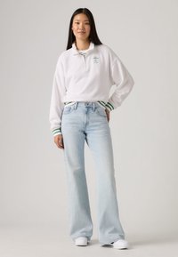 White cotton pullover with green striped cuffs and embroidery, paired with light blue flared jeans and white sneakers.