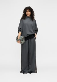 Σκούρο γκρι φαρδύ φούτερ με ψηλό γιακά και μακριά μανίκια, σε συνδυασμό με άνετο παντελόνι, κρατώντας μια τσάντα με υφή.