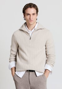 Maglione beige lavorato a maglia con collo a mezza zip, motivo strutturato e maniche a coste, indossato sopra una camicia bianca con bottoni.
