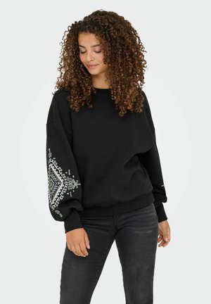 Felpa nera in morbido tessuto, con un design testurizzato bianco sulla manica sinistra. Polsini e orlo a costine. Abbinata a jeans scuri.