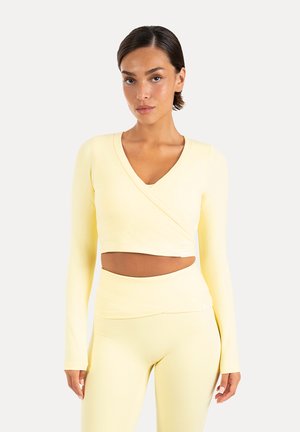 Vrouw met een lichtgele lange mouwen wikkel-crop top en bijpassende hoog getailleerde aansluitende broek, staand tegen een effen achtergrond.