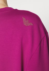 Magenta sweter dzianinowy z dużym, teksturowanym logo z złotych ćwieków na prawym ramieniu. Prosty, luźny fason.