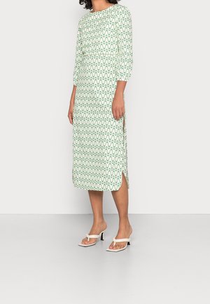 Frau trägt ein weißes Midikleid mit grünen wellenförmigen Linienmustern und Dreiviertelärmeln, kombiniert mit weißen offenen Sandaletten mit Absatz.