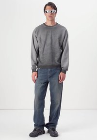 Grå sweatshirt med tekstureret ørnedesign, parret med slidte blå jeans og sorte sko. Model iført reflekterende solbriller.