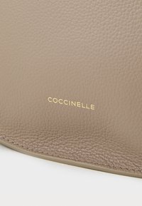 Bežinė odos paviršius su akmuo imituojančia tekstūra, puikuojantis auksiniu "Coccinelle" logotipu. Matomi lygūs kraštai ir smulkūs siuvimo detalės.