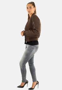 Donna in posizione laterale che indossa una giacca bomber marrone, un top nero, jeans skinny grigi sbiaditi e scarpe con tacco nere su uno sfondo bianco.